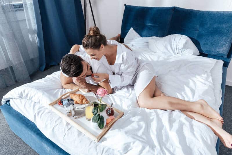 Couple smiling and hugging while lying in bed with breakfast beside them on a tray.