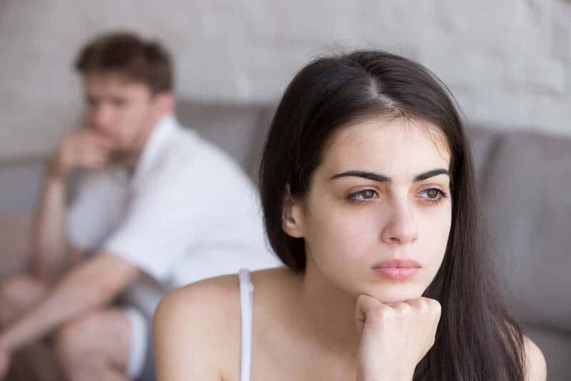 Frustrated young woman looking into the distance, thinking about her relationship. Boyfriend in background looking upset