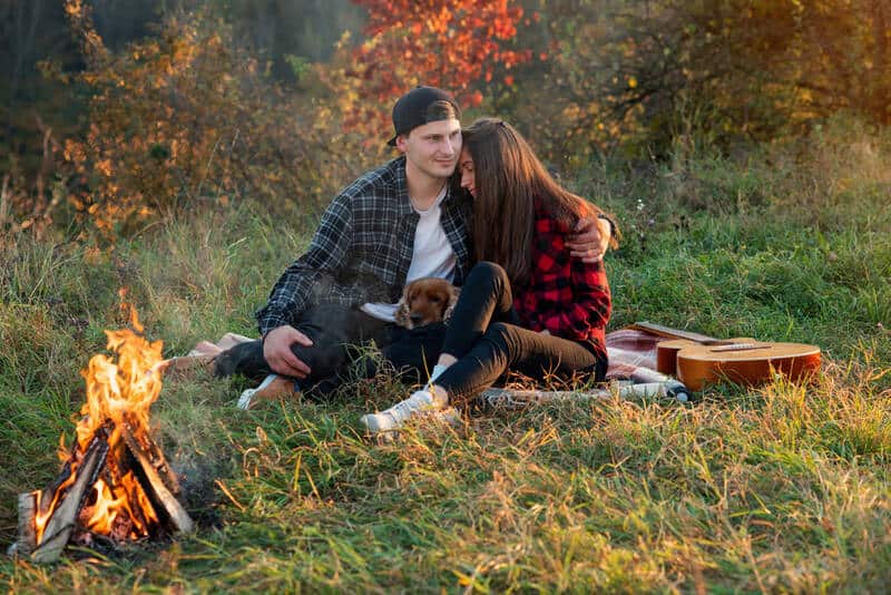 Couple dressed in casual clothes with their dog sitting in their backyard with a campfire.