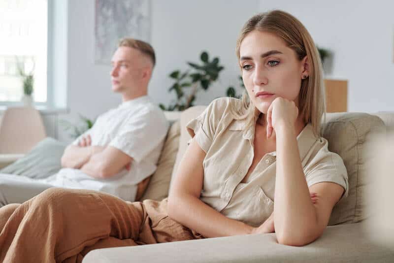 Young sad female and her offended boyfriend in background sitting on couch in living-room and keeping silence after or during argument. 